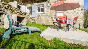 A stone building with a wooden balcony, a concrete patio with a table and chairs, and a green lounge chair. - Le Gîte de Pount d'Iher - Proche GR10 (Laruns)