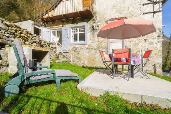 Un bâtiment en pierre doté d’un balcon en bois, une terrasse en béton avec une table et des chaises, et un fauteuil de jardin vert.