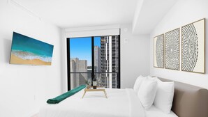Ein modernes Hotelzimmer mit einem großen Bett, Blick auf die Stadt durch raumhohe Fenster, einem Strandbild und dekorativen Wandbildern.
