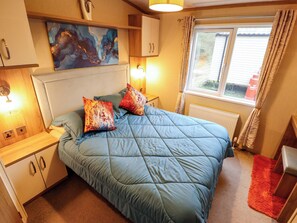 A bedroom with a bed, pillows, a nightstand, a lamp, a window with curtains, and a painting on the wall. - 108 Fox Burrow Hang (Great Yarmouth)