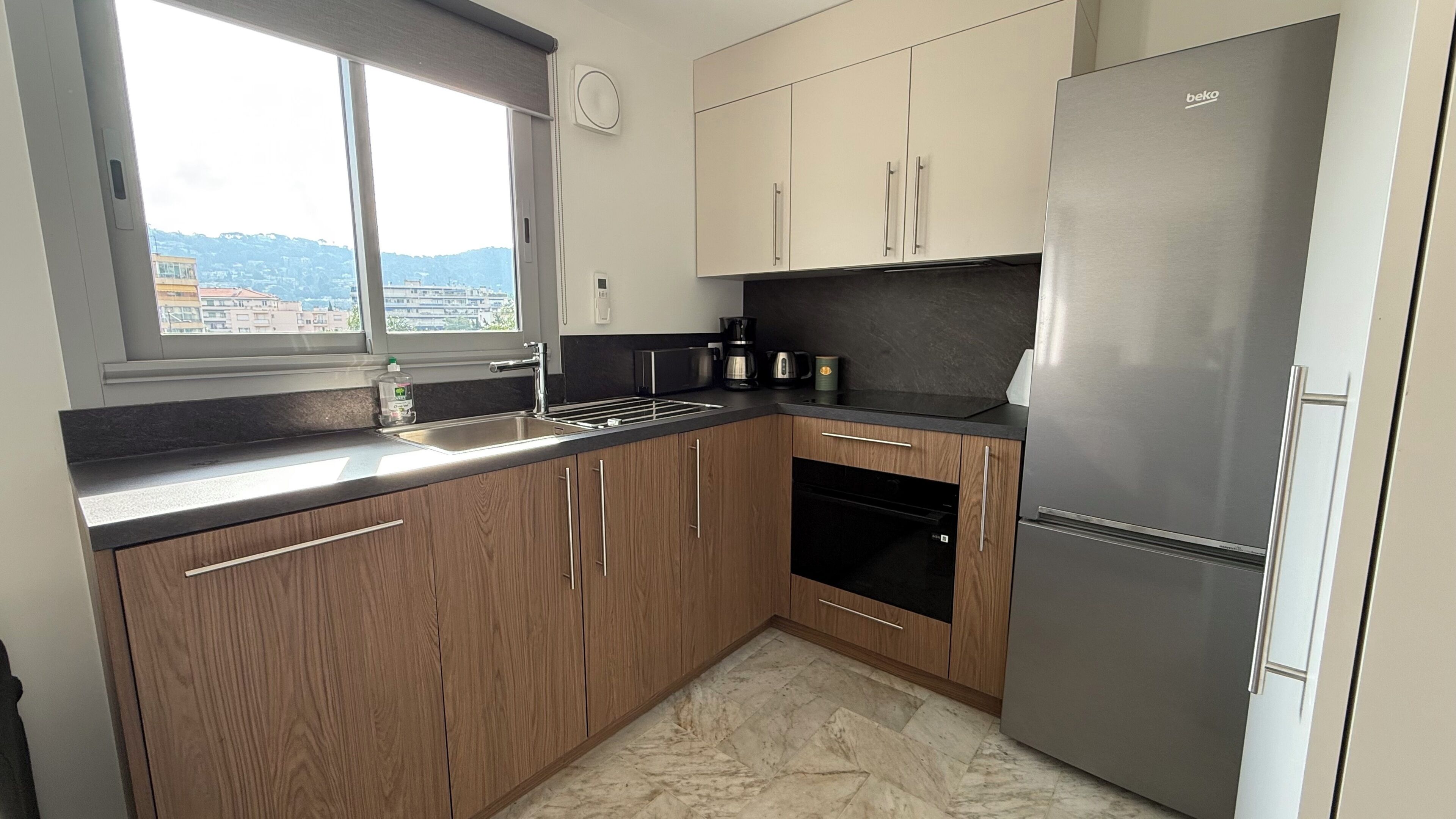 A kitchen with wooden cabinets, a stainless steel refrigerator, a coffee maker, and a window with a view of buildings.