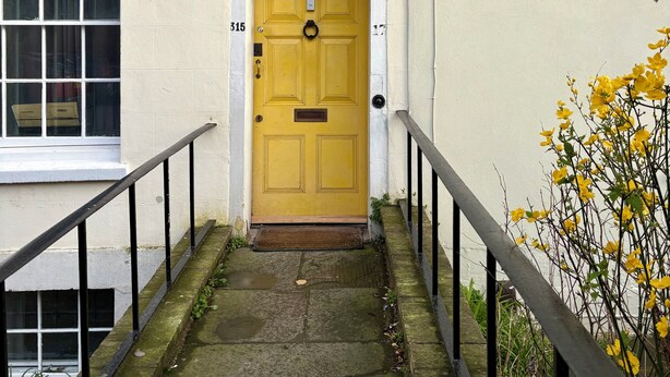 A yellow door with a small window above it, a white wall, and a black metal railing beside a stone pathway. - Home in Hotwells (Bristol)
