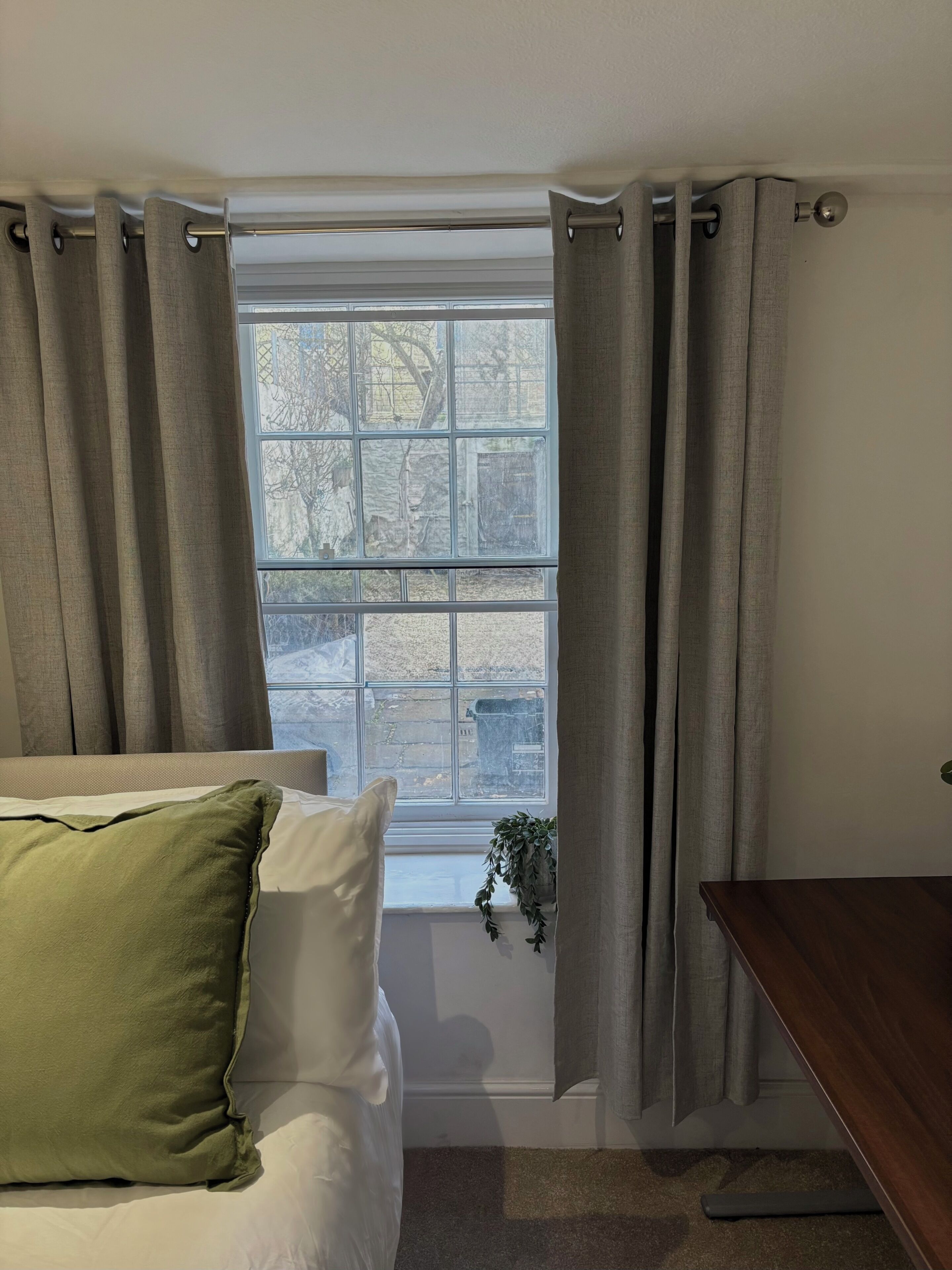 Ein Schlafzimmer mit Bett, Nachttisch, Fenster mit Vorhängen und einer Pflanze am Fensterbrett.