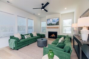 A modern living room with green sofas, a fireplace, and a ceiling fan. - 817 Cheehaw | Luxury Beach House | Sleeps 13 | Elevator | 1,000 ft to Beach Access | Dogs Welcome (Edisto Beach)