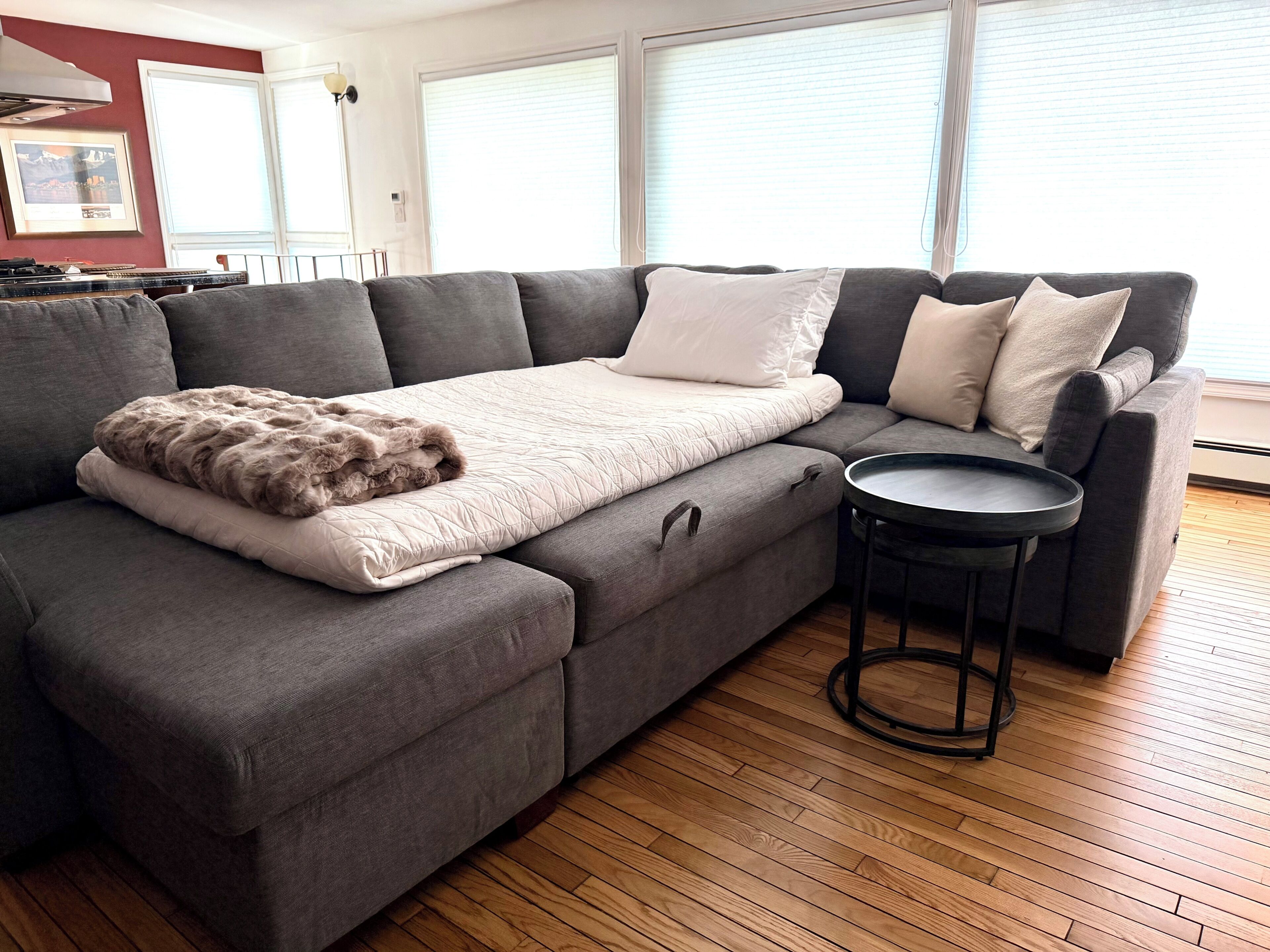 A modern living room with a grey sectional sofa, a small round table, and a view of the city through large windows.