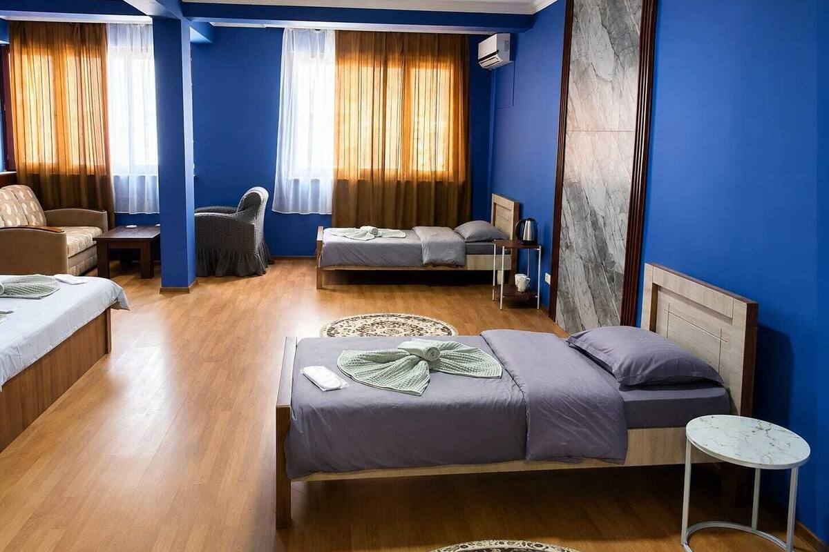 Una habitación de hotel con piso de madera, una cama con cubrecama gris y morado, un sofá, una silla, una mesita pequeña y un ventanal grande con cortinas.