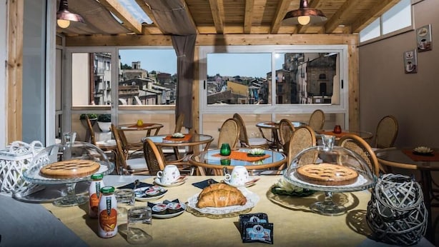 A cozy dining area with wooden beams, large windows with city views, wicker chairs, and a table set with food and drinks. - Aristonsuite (Piazza Armerina)