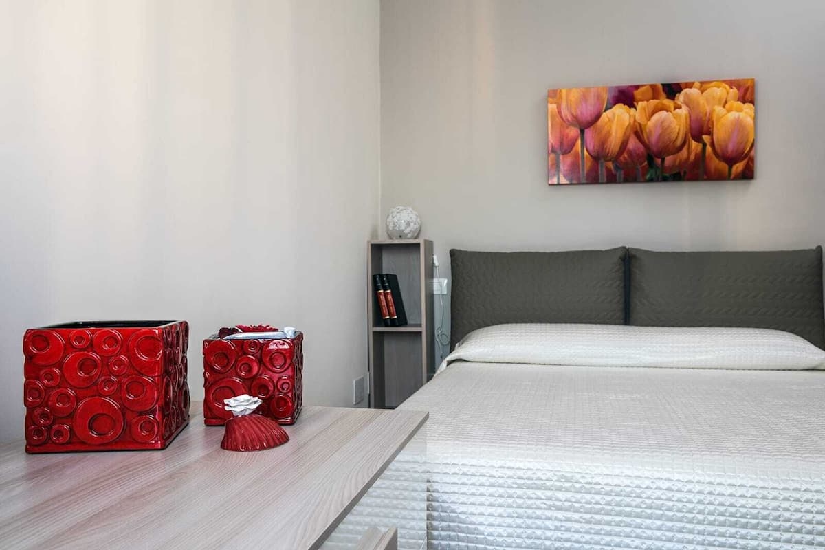 Un dormitorio con cama, una mesita de noche, una caja decorativa roja, un almohadón blanco y un cuadro con flores en la pared.