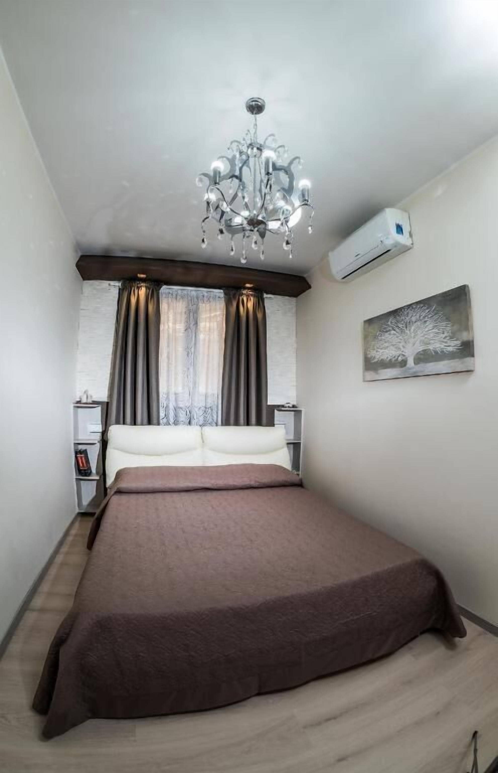 A bedroom with a bed, a chandelier, an air conditioning unit, a window with curtains, and a painting on the wall.