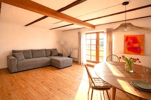 A living room with a grey sofa, wooden dining table, and chairs. There is a window with curtains, a radiator, and a hanging light fixture. - Auszeit vom Alltag: Ferienhaus zwischen Ostsee & Schlei - 4 Personen (Rieseby)