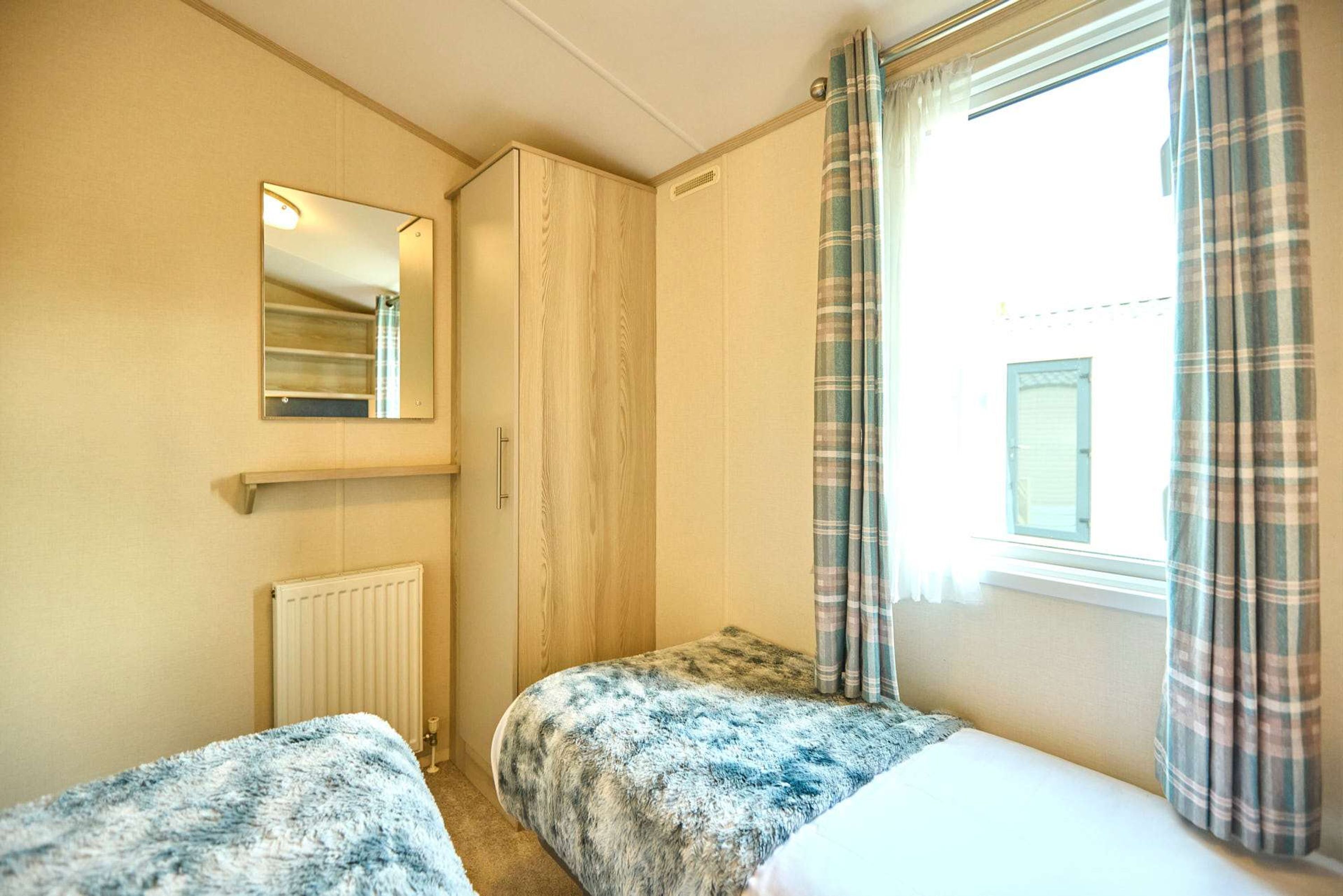 A small, single-room accommodation with a bed, a wardrobe, a mirror, and a window with curtains.