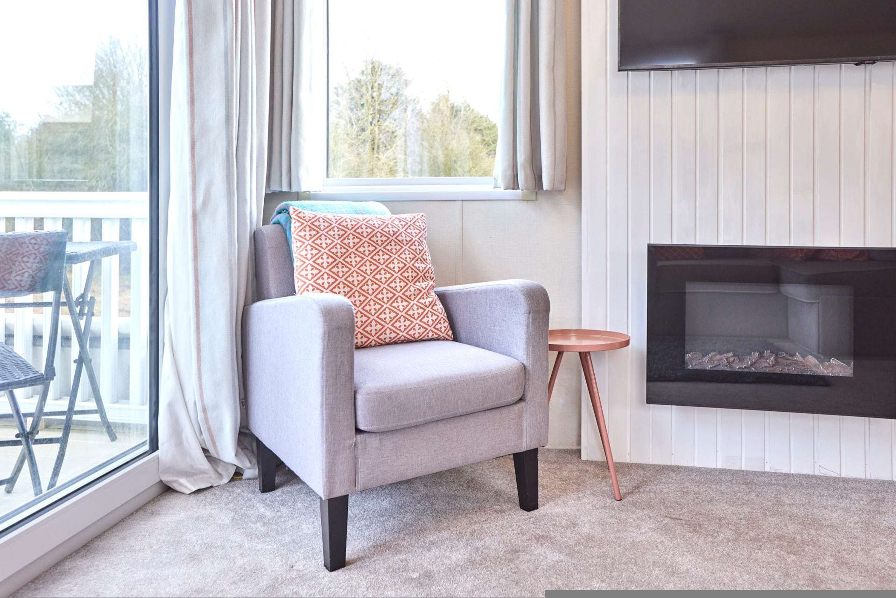 A modern living room with a grey armchair, a small round table, a fireplace, and a balcony with outdoor furniture.