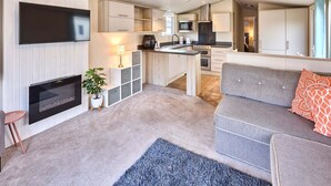 Ein modernes Wohnzimmer mit Fernseher, Kamin, grauem Sofa und einer Küchenzeile mit Schränken und Geräten.