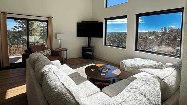 A modern living room with a sofa set, a coffee table, a flat-screen TV, and large windows offering a view of a forested area. - The Overlook on Mountain View (Lead)