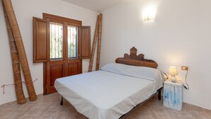 A bedroom with a wooden bed, a nightstand with a lamp, a window with wooden shutters, and bamboo poles leaning against the wall. - Holiday Home 'San Teodoro House with Garden' with Mountain View, Wi-Fi and Air Conditioning (Suaredda-traversa)