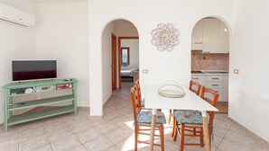 A dining area with a table and chairs, a television on a stand, and a kitchen in the background. - Holiday Home 'San Teodoro House with Garden' with Mountain View, Wi-Fi and Air Conditioning (Suaredda-traversa)