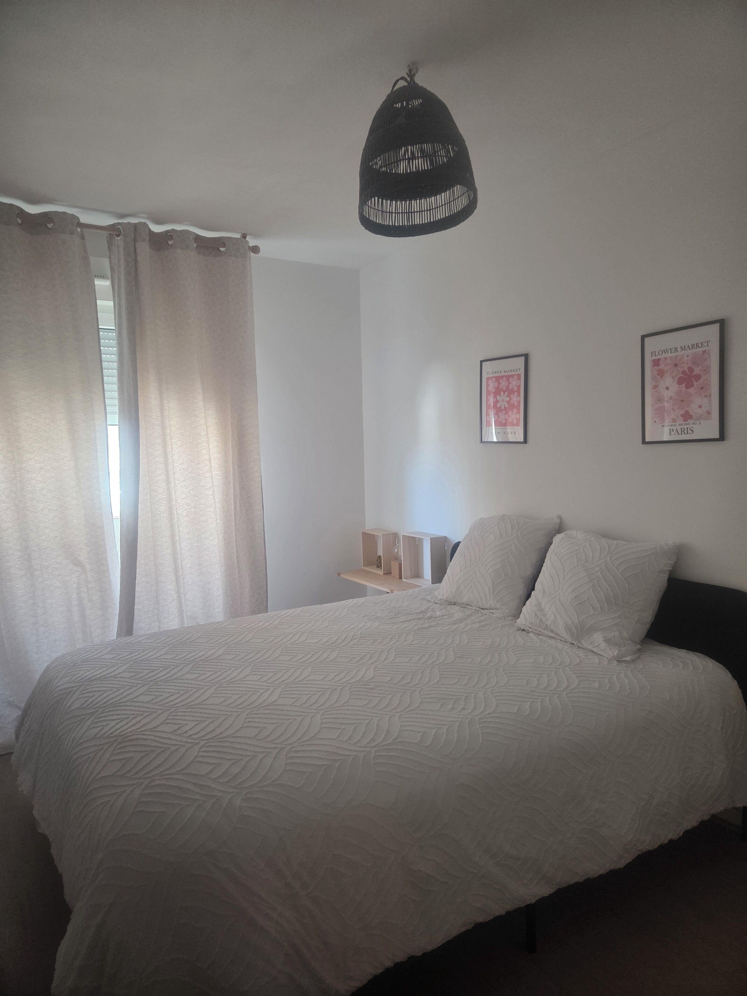 Een slaapkamer met een bed, een hanglamp, een raam met gordijnen en twee aan de muur bevestigde afbeeldingen.