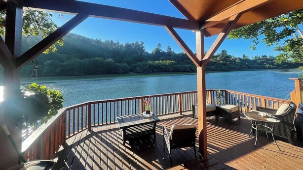 A wooden deck with a table and chairs, overlooking a lake with trees in the background. - Coastal Cabin Getaway & Animal Sanctuary (Brookings)