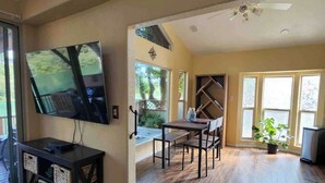 A spacious living room with a large TV, wooden flooring, a dining area, and large windows. - Coastal Cabin Getaway & Animal Sanctuary (Brookings)