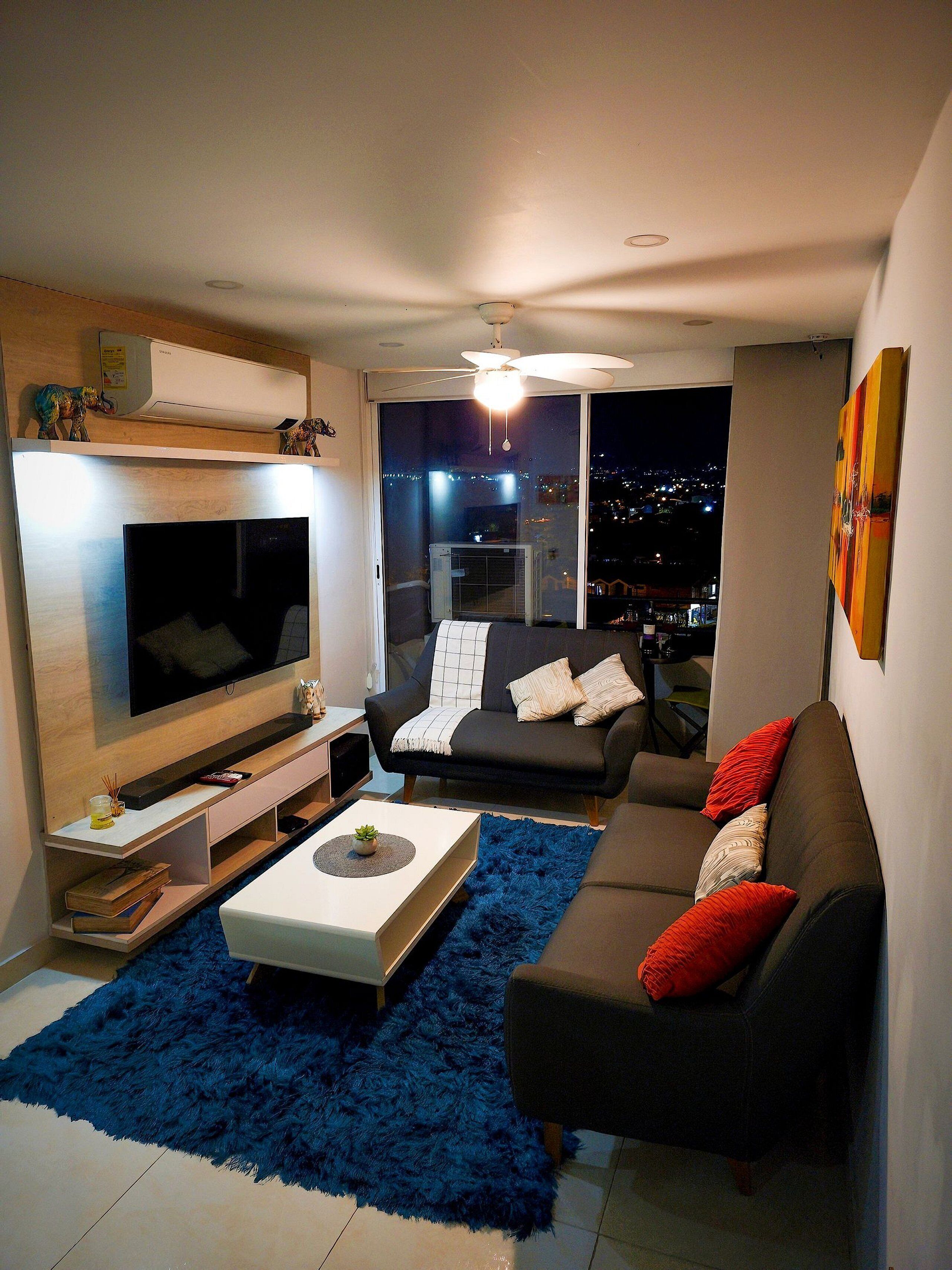 Un moderno salón con televisor de pantalla plana, un sofá negro con cojines rojos, un alfombra de pelo largo azul y vistas a la ciudad a través de la ventana.
