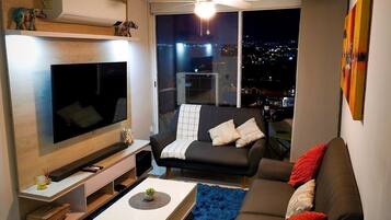 Un moderno salón con televisor de pantalla plana, un sofá negro con cojines rojos, un alfombra de pelo largo azul y vistas a la ciudad a través de la ventana.