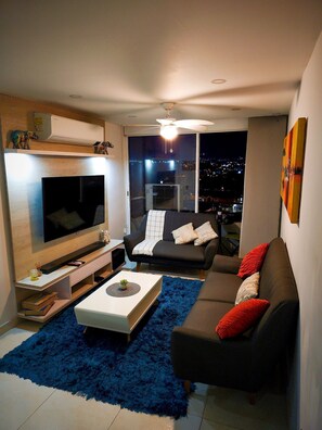 Uma sala de estar moderna com uma televisão de tela plana, um sofá preto com almofadas vermelhas, um tapete felpudo azul e vista para a cidade pela janela.