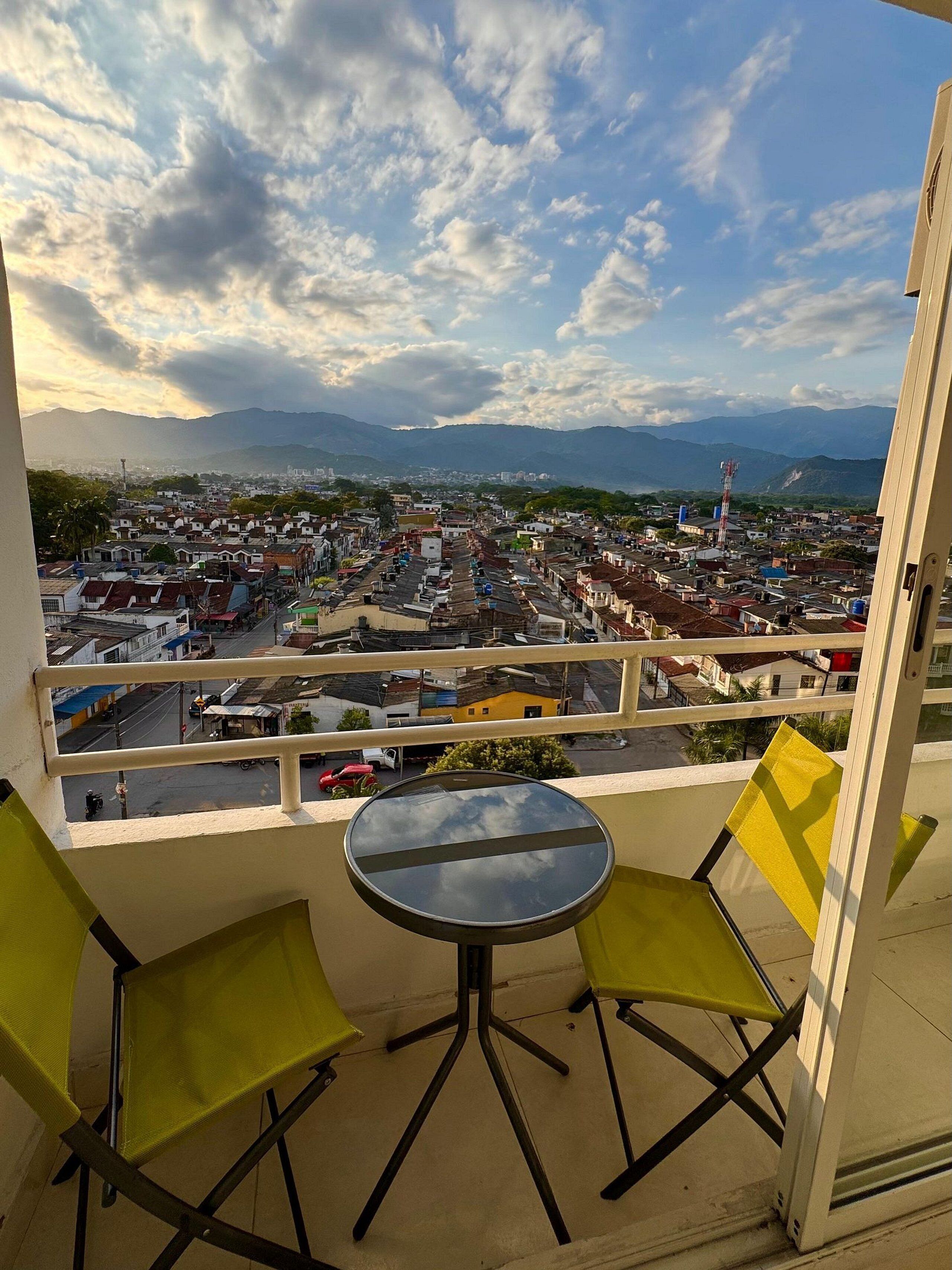 A balcony with a table and two chairs, overlooking a cityscape with buildings and mountains.