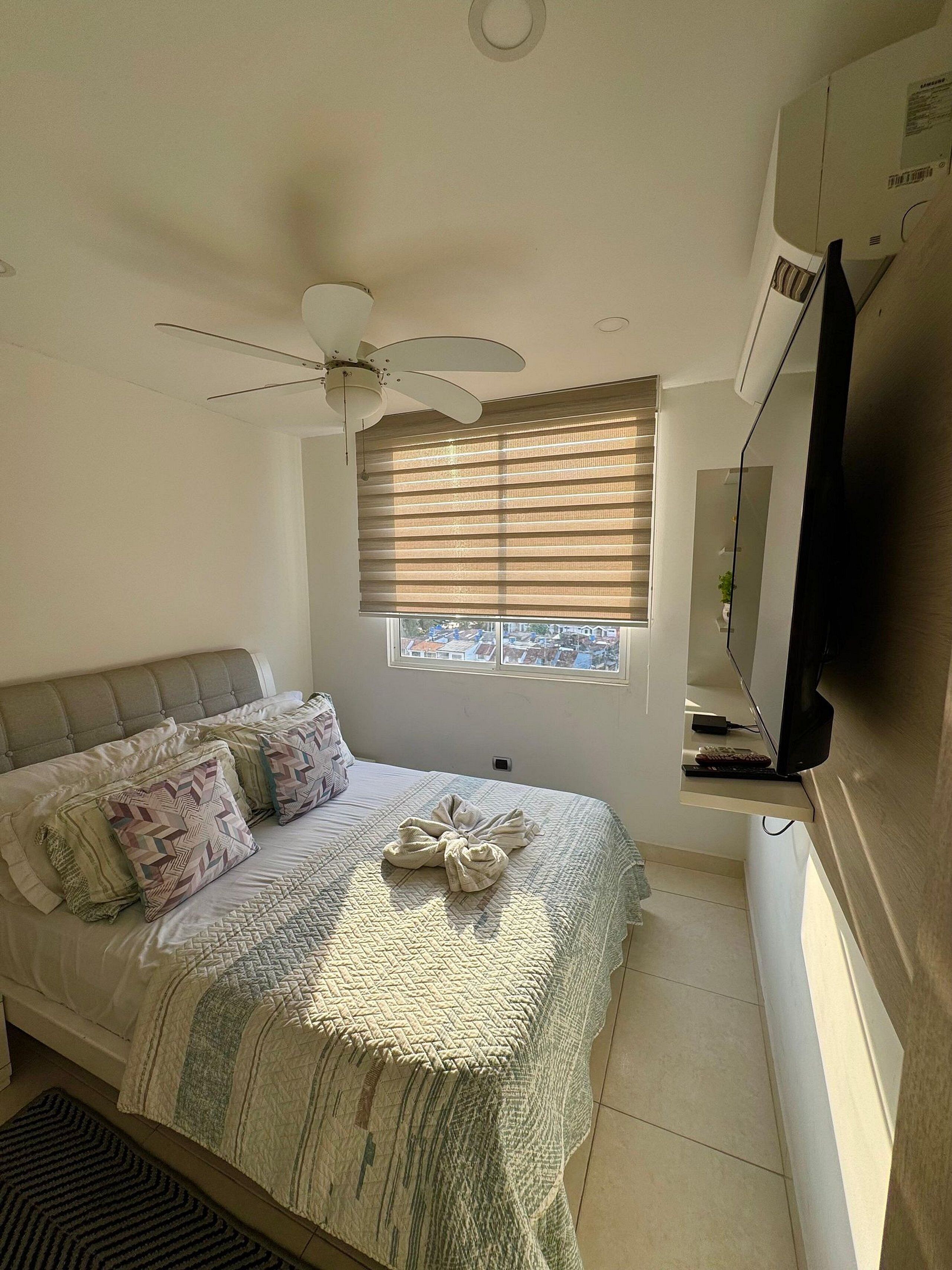 A bedroom with a bed, a ceiling fan, a window with blinds, a television, and a view of buildings outside.