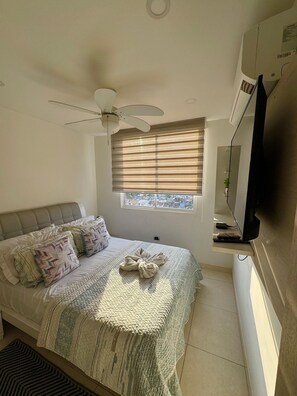 Quarto com cama, ventilador de teto, janela com persianas, televisão e vista para edifícios.