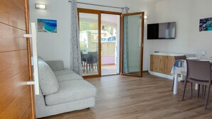 Een moderne woonkamer met een bank, een eethoek met een tafel en stoelen, een aan de muur bevestigde televisie, en een schuifdeur die leidt naar een buitenruimte.