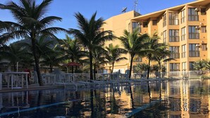 En swimmingpool med klart vand, omgivet af palmer og en bygning med altaner.