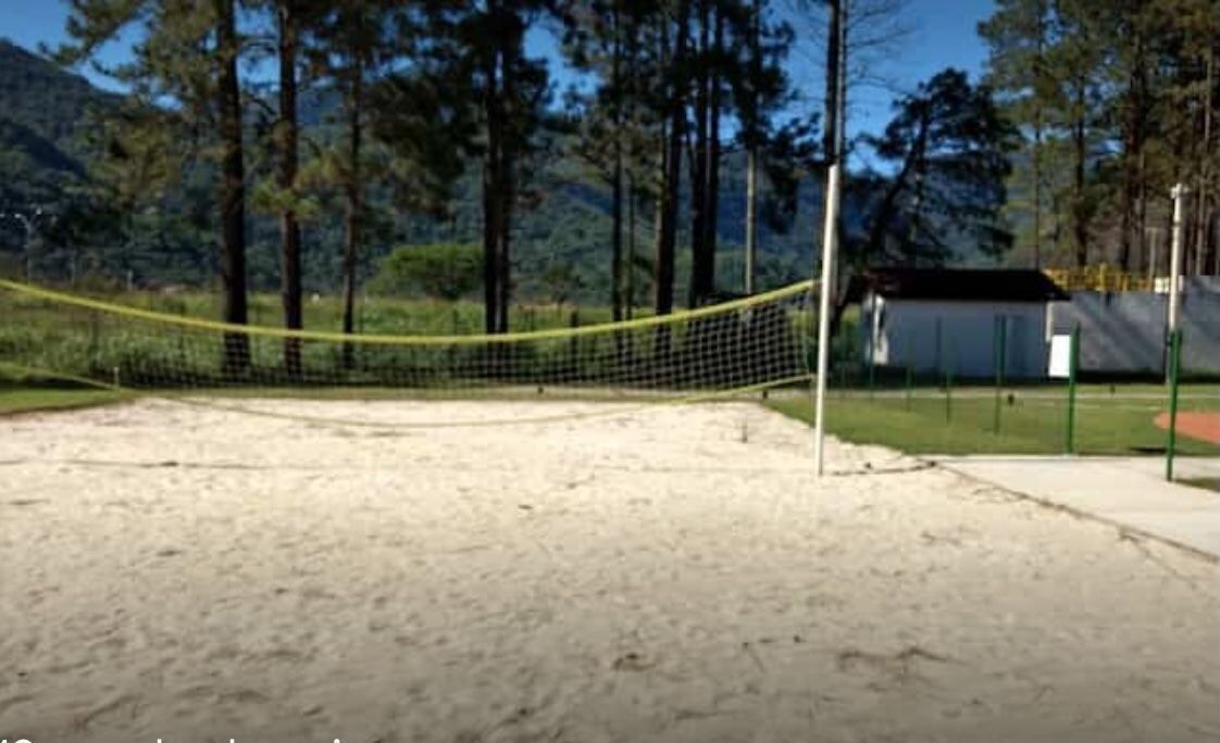 Un terrain de volleyball de sable avec un filet, entouré d’arbres et d’un petit bâtiment.