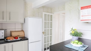 A kitchen with white cabinets, a refrigerator, a wooden cutting board with vegetables, and a bowl of apples on the counter. - Bodlasan Groes Cottage (Holyhead)