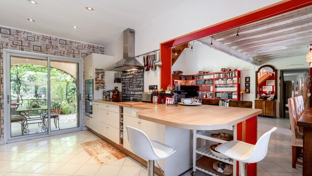 A modern kitchen with a central island, white bar stools, and a view of the garden through glass doors.
