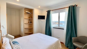 Ein Schlafzimmer mit einem Bett, einem Sessel, einem Fernseher, einem Fenster mit Vorhängen und einem hölzernen Regal.