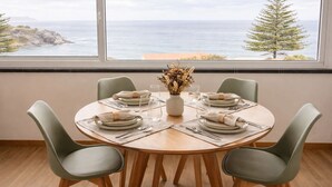 Ein runder Esstisch mit vier Stühlen, eine Vase mit getrockneten Blumen und Blick auf das Meer durch große Fenster.