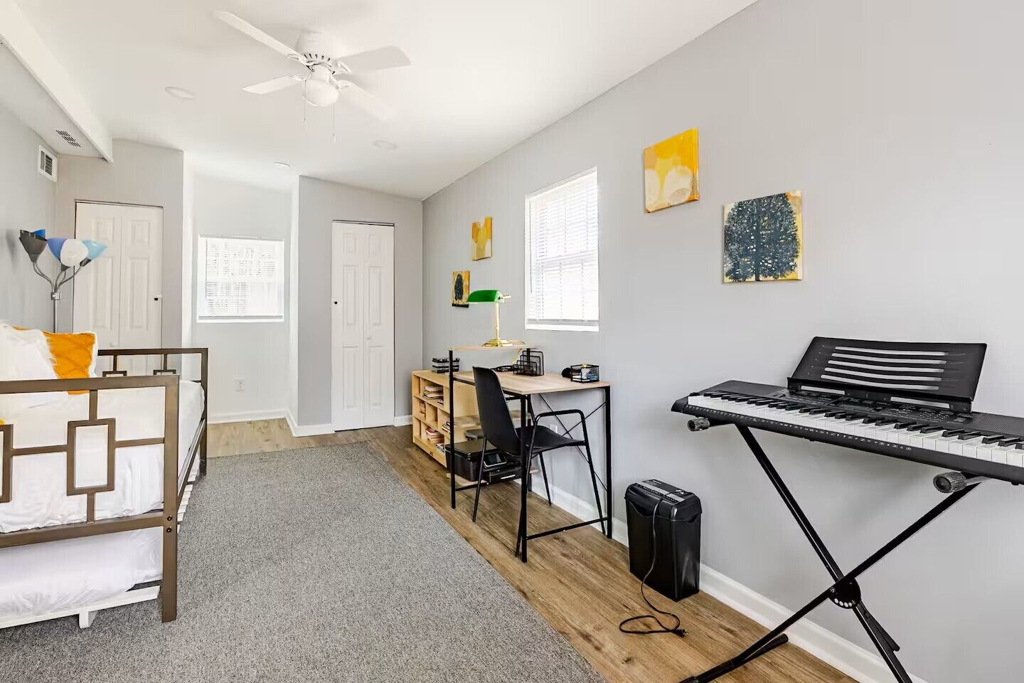 A bedroom with a bed, a desk, a chair, a standing keyboard, and a ceiling fan.