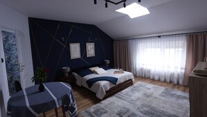Quarto com cama, mesas de cabeceira, escrivaninha, cadeira, janela com cortinas e claraboia.