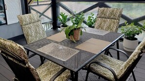 Uma área de estar coberta ao ar livre, com mesa e cadeiras, rodeada por plantas em vasos e vista para o verde.