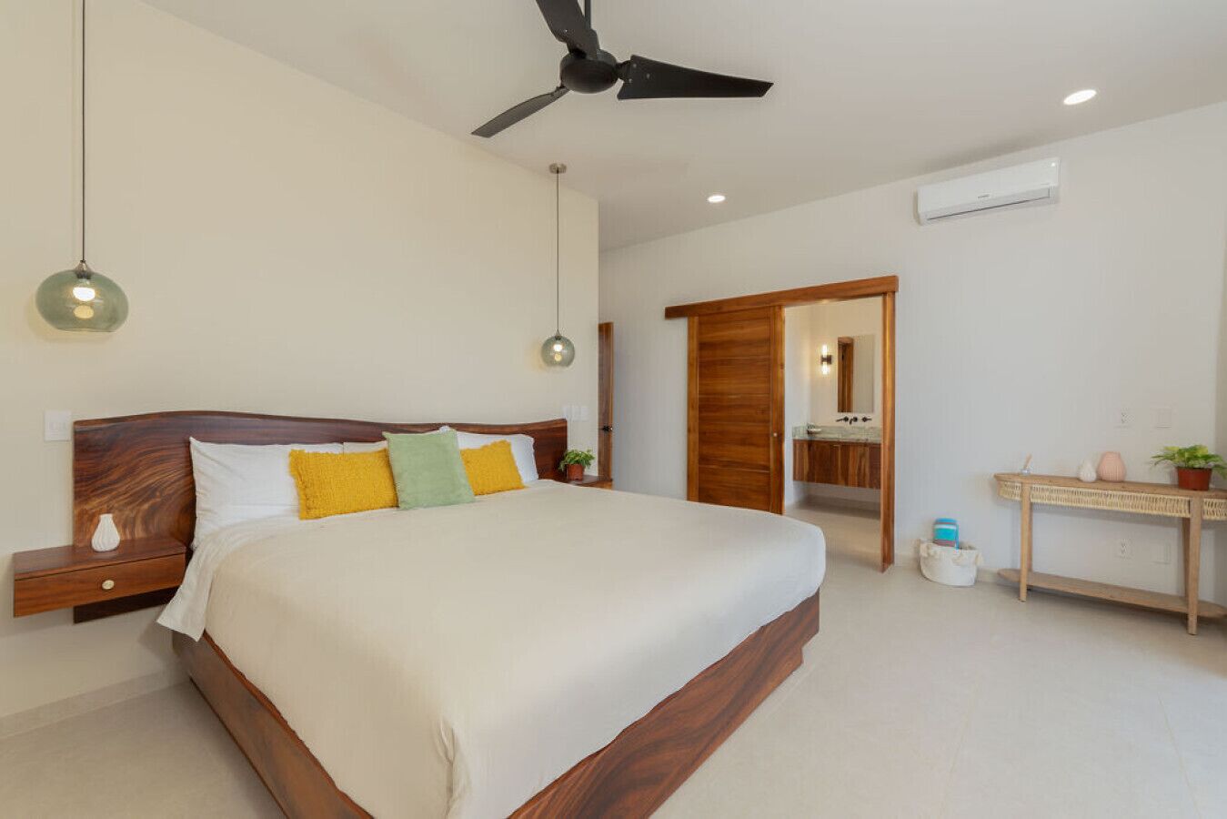 Un dormitorio con cama, mesitas de noche, ventilador de techo, una lámpara, puerta de madera y un baño visible a través de un vano abierto, junto con una pequeña mesa con una planta.