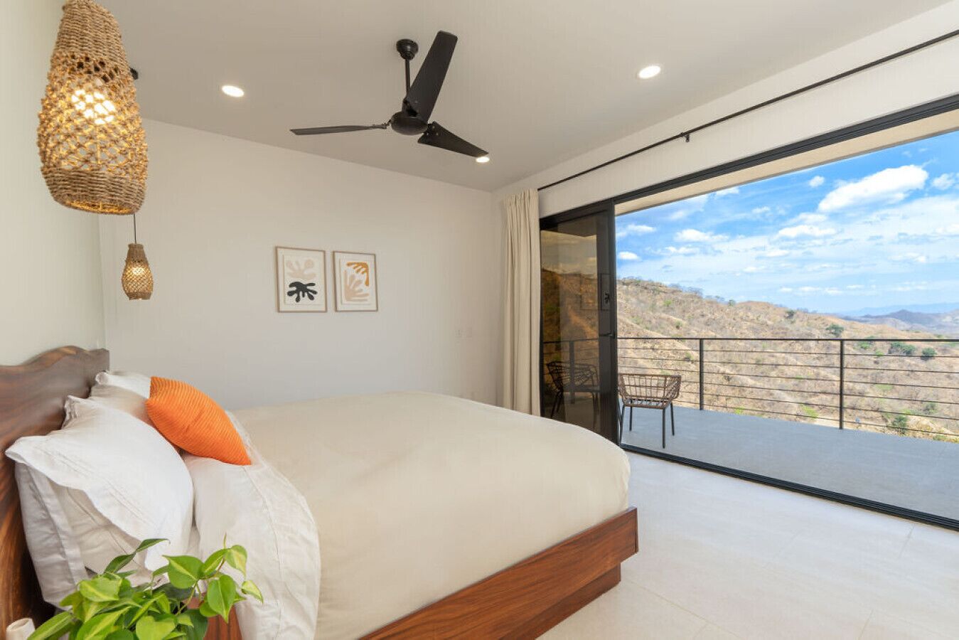 A bedroom with a bed, a ceiling fan, a balcony with a view of the hills, and two framed pictures on the wall.