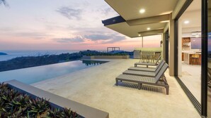 Un área de piscina moderna al aire libre con sillones de descanso, una parrilla y vista al océano al atardecer.