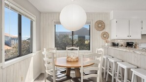 Uma sala de jantar com uma mesa redonda, cadeiras brancas, um lustre e vista através de grandes janelas.