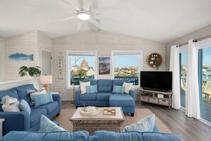 A living room with a blue sofa set, a flat-screen TV, and a large window with a view of the sea. - King Tide Rising by Rather Be Properties (Avon)