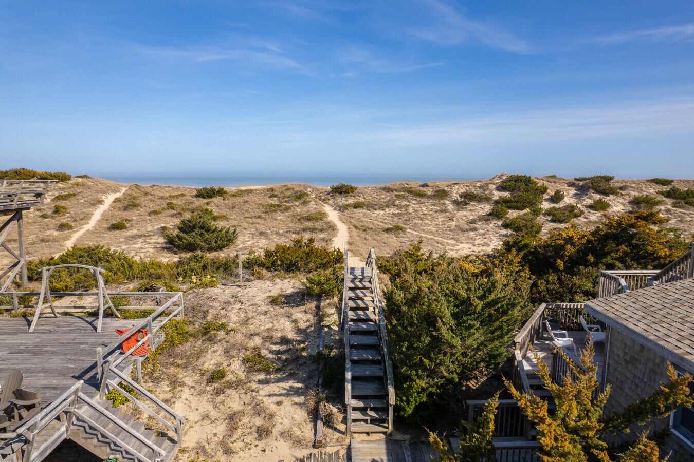 Vista costera con un paseo marítimo de madera que conduce a una playa arenosa, rodeada de vegetación escasa y un cielo despejado.