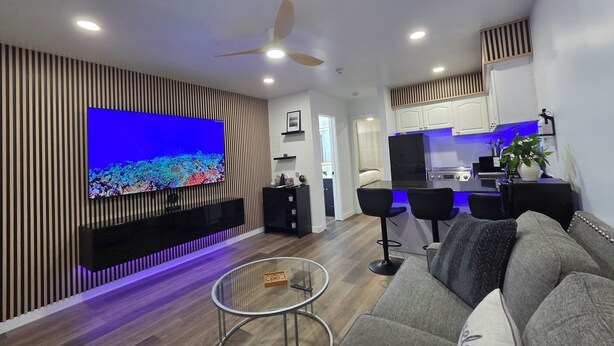 Una sala de estar moderna con un televisor de pantalla plana, un ventilador de techo y una cocina con armarios blancos y un refrigerador negro.
