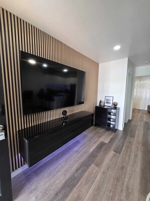 A modern living room with a large flat-screen TV mounted on a wooden panel wall, a black entertainment unit, and a patterned wooden floor.
