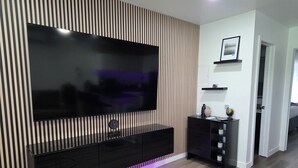 Una sala de estar moderna con un televisor de pantalla plana grande, un diseño de pared de listones de madera, un mueble de entretenimiento negro y una puerta que conduce a otra habitación.