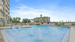 A swimming pool with multiple fountains, a poolside area with lounge chairs, and a building with a green roof in the background. - Beachside II #4247 (Miramar Beach)