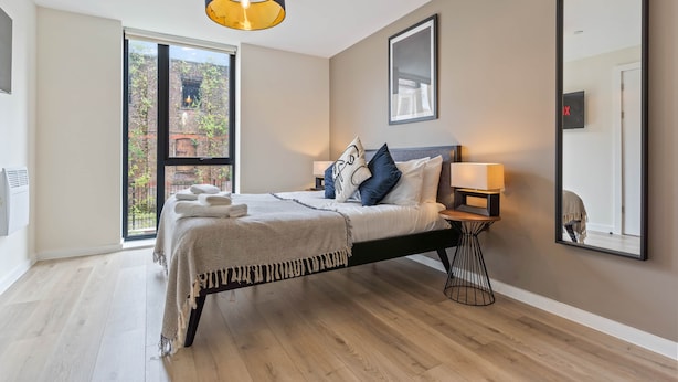 A bedroom with a bed, bedside tables, a hanging light fixture, a large window, and a mirror on the wall. - Liverpool Richmond Row 2 Bed/2 Bath Flat (107)| Secure Parking, WiFi & EV (Liverpool)